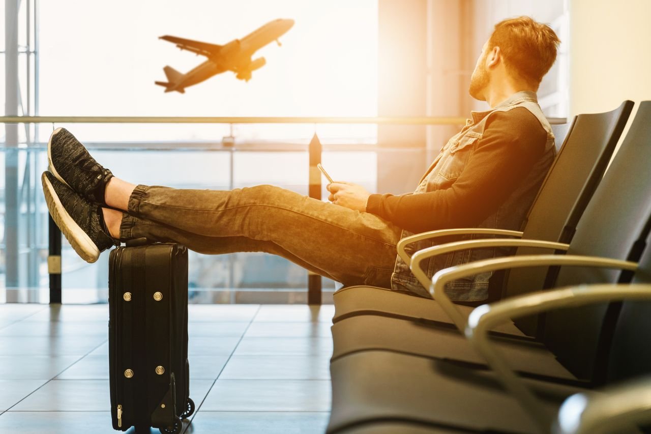 gallery-01 Side view full body of bearded man sitting on chair in waiting hall of airport and looking at flying aircraft behind window