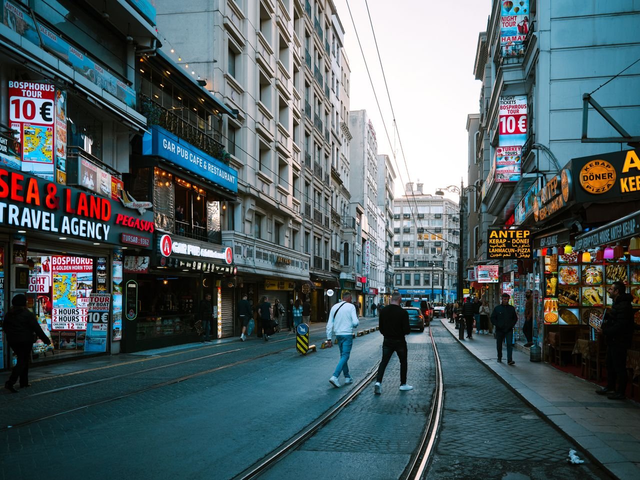 about-img-01 Vibrant street scene in Istanbul featuring shops, people, and tram tracks under a clear sky.