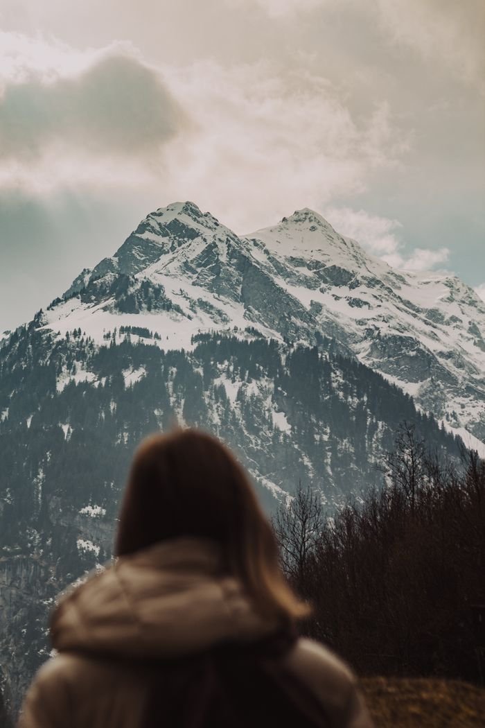 services-01 A woman in a coat looks at a majestic snow-capped mountain under cloudy skies.
