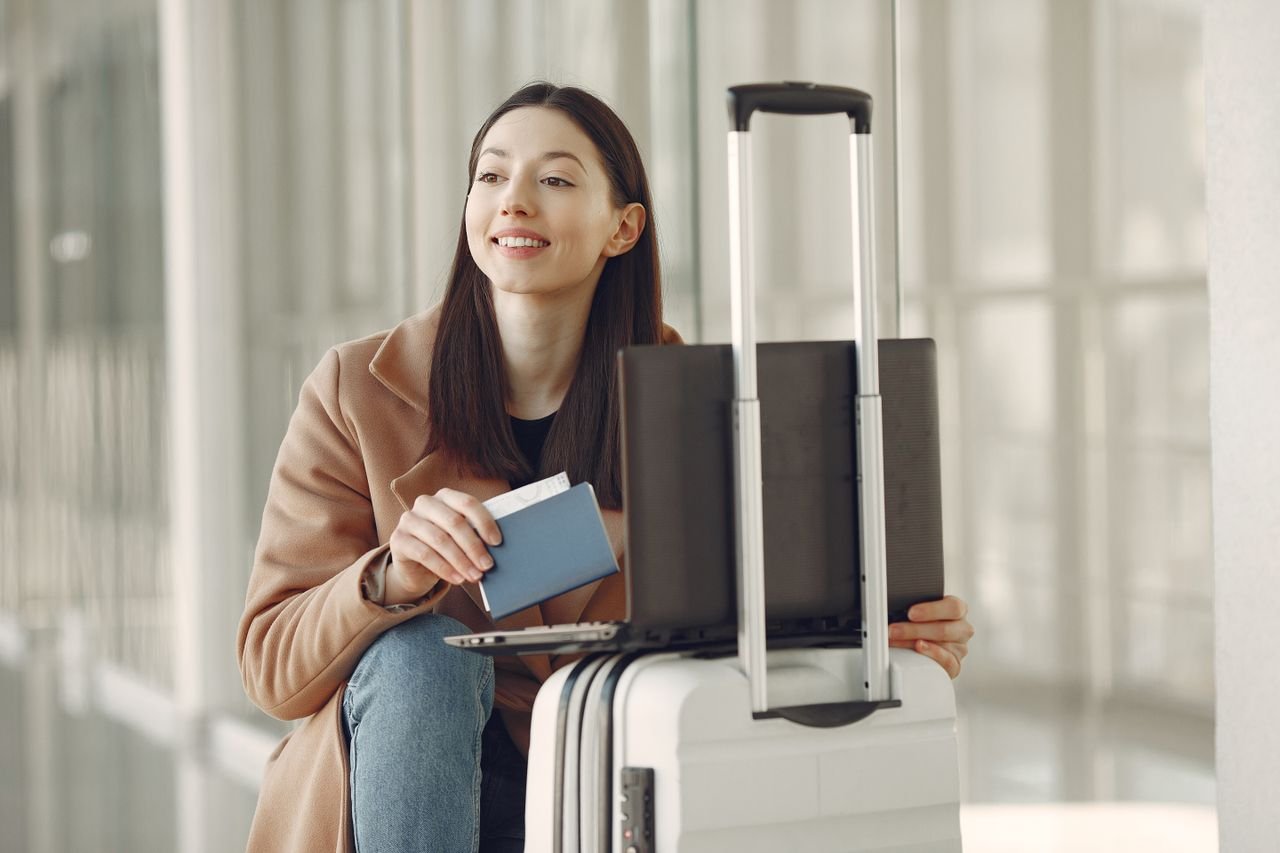 Smiling adult ethnic female traveler in trendy coat holding passport and using laptop on luggage in airport corridor for checking ticket for correctness
