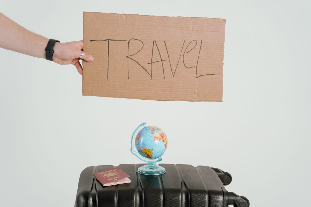 home-img A hand holds a travel sign above a globe and suitcase, symbolizing wanderlust.